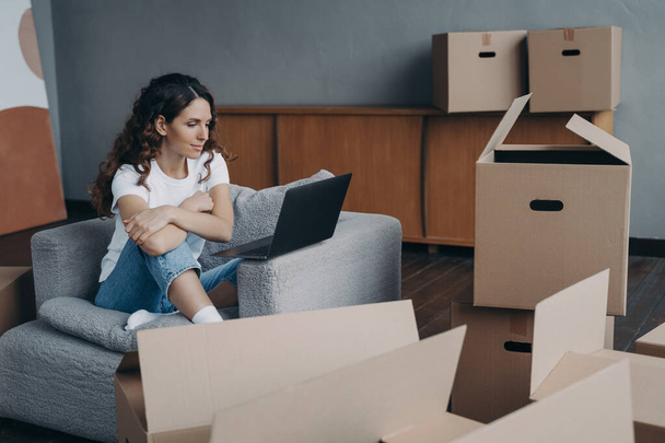 Happy young european woman selling apartment through internet. Girl among cardboard boxes is using laptop and smiling. Lady is packing things. Relocation concept. - Φωτογραφία, εικόνα