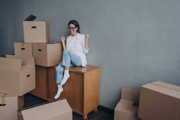 Overjoyed young woman purchases real estate. Hispanic girl is sitting among cardboard boxes in new house and celebrating. Concept of success and opportunities for women. Investment and mortgage. - 写真・画像