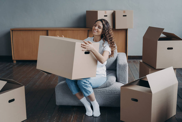 Proud european girl relocates to new home. Happy young woman moves alone. Lady is sitting in armchair among packed cardboard boxes. Independence, life choice and success concept. - Photo, Image