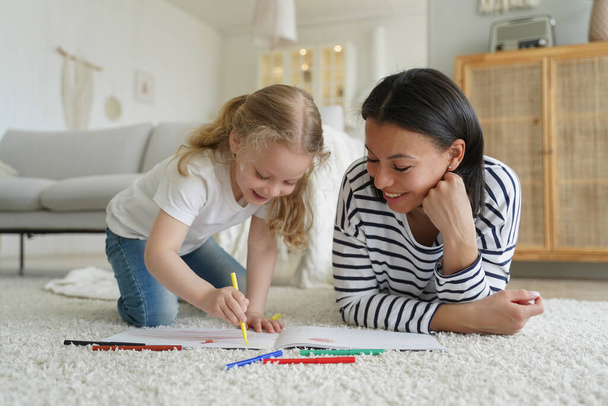 Uśmiechnięta matka i mała adoptowana córka malują razem, leżąc na podłodze. Mama lub opiekunka pomaga przedszkolance rysować ołówkami. Szczęśliwego macierzyństwa, koncepcji adopcji. - Zdjęcie, obraz
