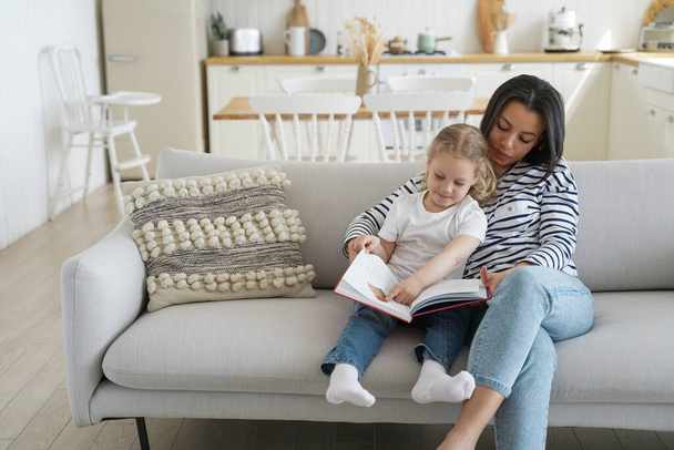 Young mom and adopted preschool daughter read book together, sitting, hugging on sofa at home. Mother educating little girl child, reading fairytale story on couch. Motherhood, adoption concept. - Foto, Imagem
