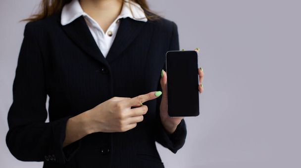 Young business woman in suit finger pointing touchin and showing blank black smartphone screen on grey background. - Photo, Image