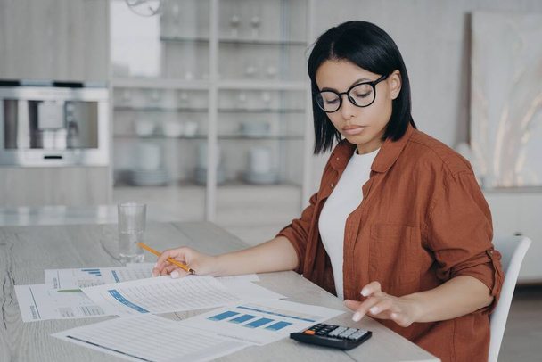 Serious girl, accountant is examining reports. Young european woman in glasses is working with data charts from home. Manager or business assistant is sitting at the desk and doing business. - Photo, Image
