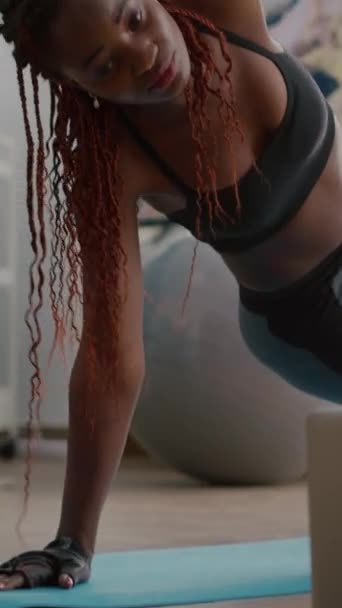 Verical video: Αθλήτρια μαύρη γυναίκα που εκπαιδεύεται στο χάρτη της γιόγκα στο σαλόνι και στέκεται σε πλαϊνή σανίδα παρακολουθώντας βίντεο γυμναστικής χρησιμοποιώντας φορητό υπολογιστή. Ενήλικες σε αθλητικά ενδύματα ραβδώσεις μυών κατά τη διάρκεια της γυμναστικής strech - Πλάνα, βίντεο