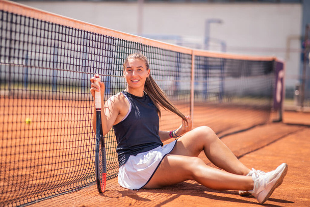 Taking video outdoor clay court charismatic with a beautiful smile professional tennis player woman holding the racket on hands and smiling large while sitting down on the floor. Portrait - Фото, зображення