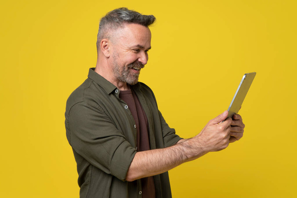 A joyful happy styled man dressed in green shirt looking into digital tablet pc holding in hands reading news or watching funny video isolated on yellow background - Zdjęcie, obraz
