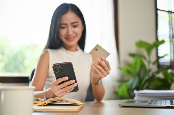 Belle femme asiatique millénaire utilisant son smartphone et sa carte de crédit pour payer ses factures en ligne. Paiement en ligne et mode de vie sans espèces concept - Photo, image
