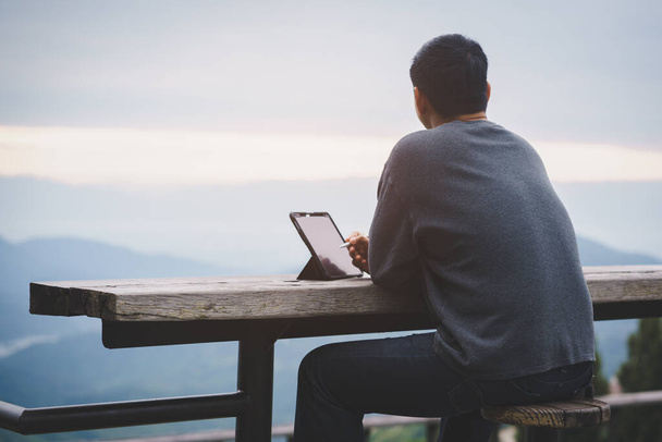 Freelancer τύπος χρησιμοποιώντας tablet φορητό υπολογιστή, ενώ κάθεται κατά ορεινό τοπίο κατά τη διάρκεια των διακοπών το καλοκαίρι ταξίδι.Έννοια του ταξιδιού και online εργασίας - Φωτογραφία, εικόνα