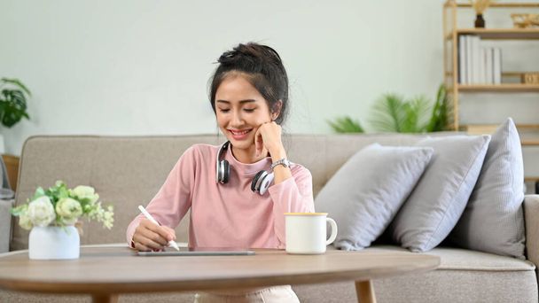 Очаровательная молодая азиатка-студентка делает домашнее задание. Женщина пишет дневник или дневник в своей гостиной. Концепция образа жизни - Фото, изображение