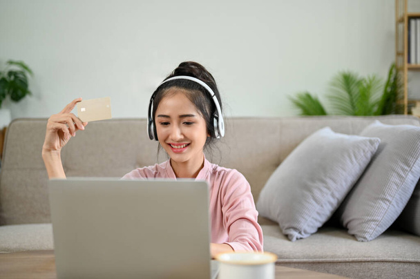 Pretty young Asian female in the living room wearing headphones, using her laptop computer, shopping online or paying bills with her credit card. - Foto, Bild