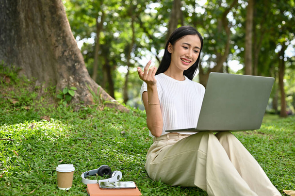 Ελκυστική νεαρή Ασιάτισσα φοιτήτρια ή γυναίκα ελεύθερη επαγγελματίας κάθεται στο γρασίδι, χρησιμοποιώντας φορητό φορητό φορητό υπολογιστή της, ενώ χαλαρώνει στο πάρκο της πόλης. - Φωτογραφία, εικόνα