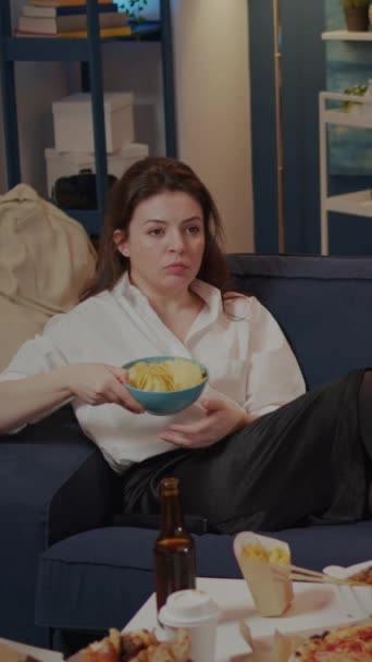 Вертикальное видео: молодая женщина лежит на диване, держа миску чипсов, смотря телевизор дома. Человек с вынос фаст-фуд на стол отдыха на диване после работы, имея перекусить в гостиной - Кадры, видео