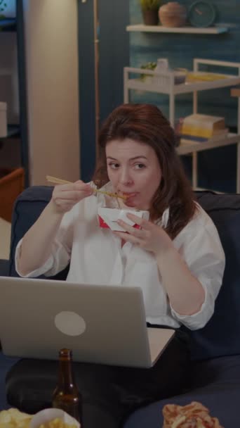 Dikey video: Koltukta dizüstü bilgisayar kullanan genç bir kadın yemek çubuklarıyla Asya yemekleri yiyor. Kafkasyalı biri evde Çin yemeği yiyor, TV 'de film seyrediyor ve cihaz üzerinde çalışıyor. - Video, Çekim
