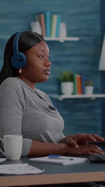 Video verticale: Studente con la pelle nera con le cuffie mette in ascolto corso universitario online utilizzando piattaforma di elearning seduto alla scrivania in soggiorno. afro americano donna lavoro remoto da casa - Filmati, video