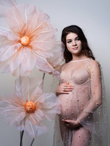 Tenera foto di una giovane donna incinta in lingerie nuda e un vestito trasparente ornato di perle, su uno sfondo leggero e con enormi fiori decorativi in piena crescita. - Foto, immagini