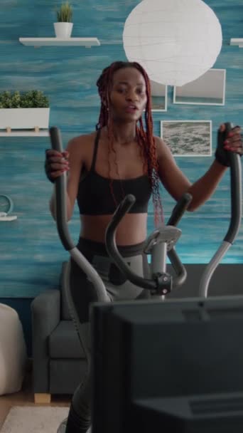 Verical video: Black woman athletic training on elliptical bike practing cardio sport παρακολουθώντας αερόβια βίντεο στην τηλεόραση κατά την πρωινή ρουτίνα σωματικού βάρους στο σαλόνι. Εκπαιδευτής απολαμβάνοντας υγιή - Πλάνα, βίντεο