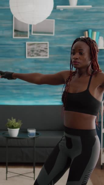 Vertikales Video: Sportliche Frau mit schwarzer Haut wärmt sich vor dem Aerobic-Training im Stehen auf einer Yogakarte im Wohnzimmer auf. Erwachsene dehnen Körpermuskeln und genießen einen gesunden Lebensstil - Filmmaterial, Video