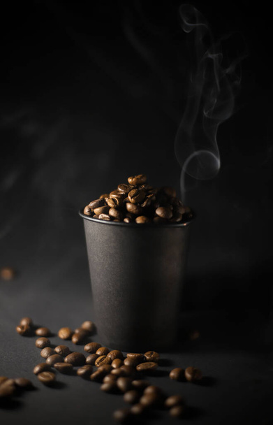 on a black background, a black craft glass full of coffee beans and brown color and white smoke and scattered grains around the glass lie nearby.for coffee houses, street menus, cafes, labels, flyers, banners - Foto, afbeelding