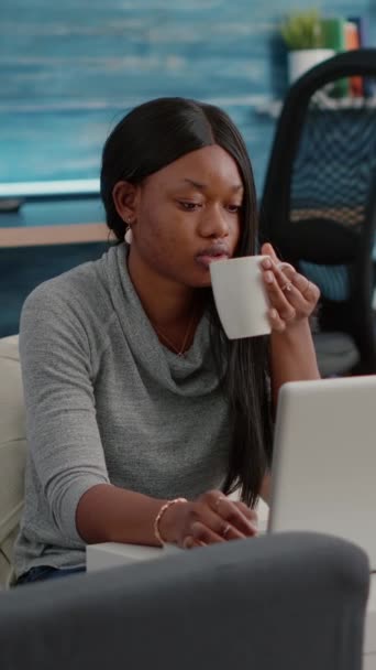 Вертикальное видео: студент блоггера с темной кожей, работающий из дома просматривая информацию связи во время питья кофе сидя на фасолью мешок в гостиной. Африканская женщина, работающая из дома в СМИ - Кадры, видео
