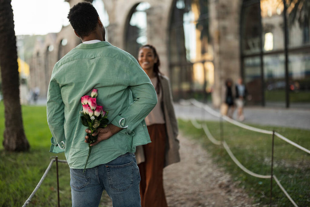 Африканский мужчина прячет за собой красные цветы, чтобы удивить свою девушку на улице. - Фото, изображение