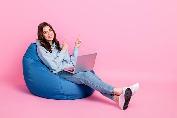 Full length portrait of attractive cheerful person sit cozy bag use netbook indicate fingers empty space isolated on pink color background. - Фото, изображение