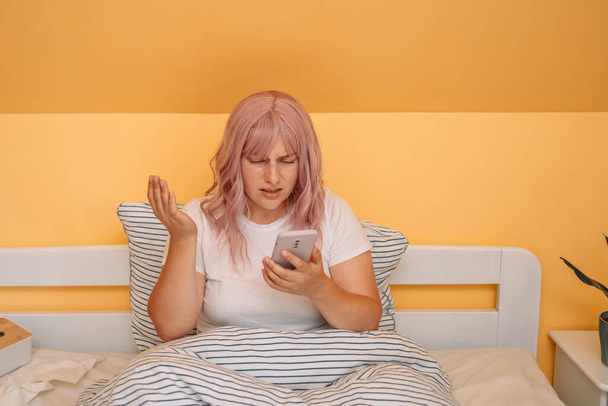 Shocked young woman waking up and looking at smartphone in bedroom at home. - Фото, изображение