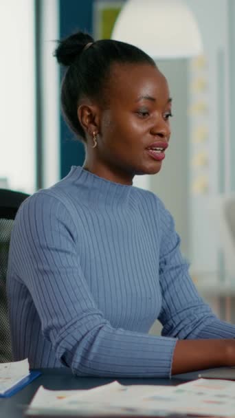 Portrait of african american woman typing using laptop keyboard and smiling at camera sitting at desk in busy startup office. Casual business employee working relaxed with sales statistics. - Video, Çekim