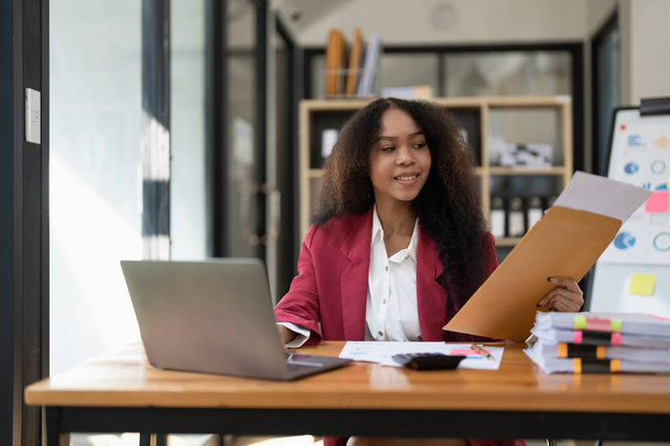 Sorridente giovane afroamericana donna Finanza Imprenditore seduto in ufficio sul posto di lavoro accumulando tazza di caffè e lavorando su un computer portatile, investimento finanziario documento relazione analisi concetto di finanza - Foto, immagini