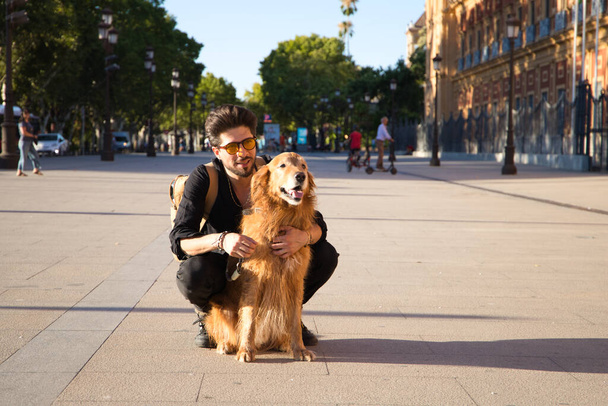 Молодий привабливий іспаномовний чоловік зі своїм коричневим золотим ретриверсійним собакою, присів униз і дивиться на те ж місце. Концепція домашніх тварин, собак, любовних тварин, золотого ретривера. - Фото, зображення