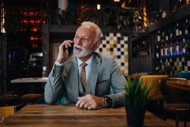 Happy businessman sitting in restaurant and waiting for lunch. He is using smart phone and talking with someone. Business seniors lifestyle concept. - Foto, imagen