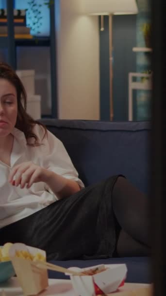 Вертикальное видео: взрослый кавказский отдыхает на диване с чипсами в качестве закуски во время еды быстрого питания от доставки на стол. Женщина, сидящая на диване и смотрящая фильм по телевизору, ест из миски в жизни - Кадры, видео