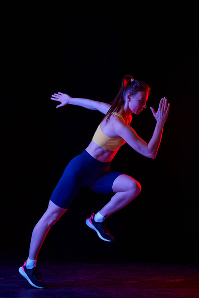 Professional runner, jogger running isolated on dark background in neon light. Healthy lifestyle, sport, fitness, speed and energy concept. Athletic woman in sports uniform and footwear training. - 写真・画像