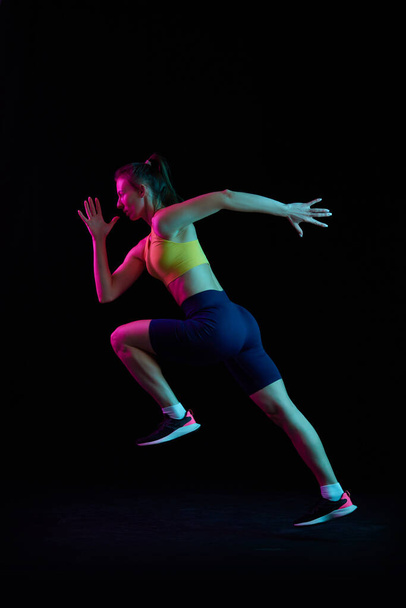 Portrait of young sportive woman training, running isolated on dark background in neon light. Competition, sport, fitness, speed and energy concept. Dynamic movements. - Φωτογραφία, εικόνα