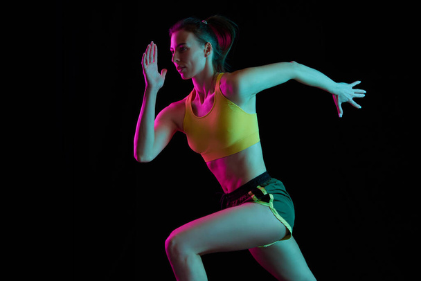 Professional runner, jogger running isolated on dark background in neon light. Healthy lifestyle, sport, fitness, speed and energy concept. Athletic woman in sports uniform and footwear training. - Φωτογραφία, εικόνα