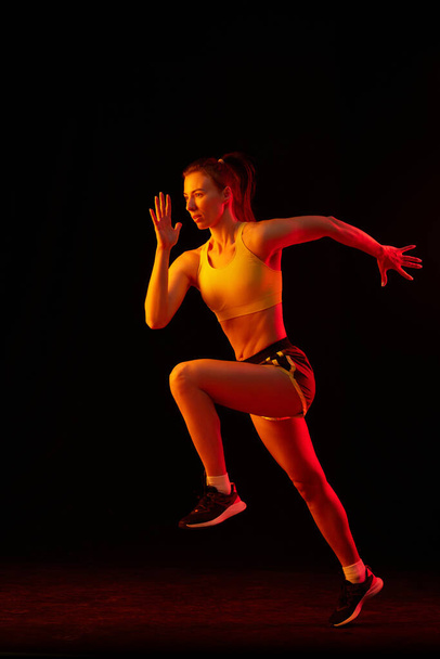 Portrait of young sportive woman training, running isolated on dark background in neon light. Competition, sport, fitness, speed and energy concept. Dynamic movements. - Foto, imagen
