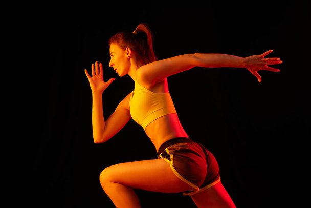 Portrait of young sportive woman training, running isolated on dark background in neon light. Competition, sport, fitness, speed and energy concept. Dynamic movements. - 写真・画像