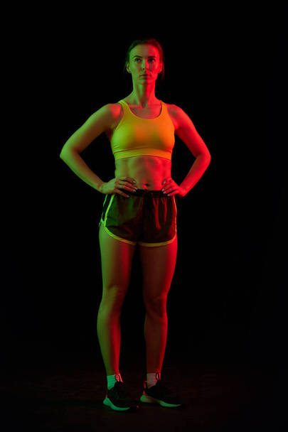 Portrait of professional female athlete posing isolated on dark background in neon ligh. Health, sport, fitness, speed and energy concept. Athletic woman in sports uniform and footwear training. - 写真・画像