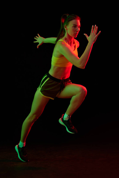 Running technique. Professional female jogger running isolated on dark background in neon light. Healthy lifestyle, sport, fitness, speed and energy concept. Sportive woman showing base movements - Photo, image