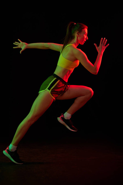 Professional runner, jogger running isolated on dark background in neon light. Healthy lifestyle, sport, fitness, speed and energy concept. Athletic woman in sports uniform and footwear training. - Photo, image