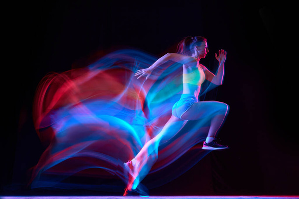 Professional female athlete, runner in motion over dark background in mixed neon light. Sportive girl in sportswear practicing in run or jogging. Healthy lifestyle concept. - Foto, Imagen