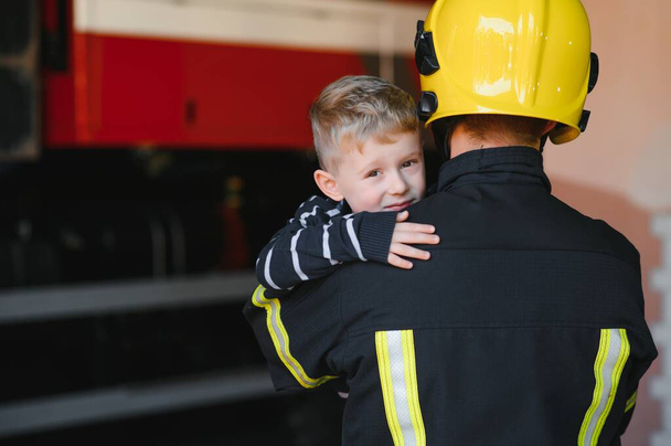 A firefighter take a little child boy to save him. Fire engine car on background. Fireman with kid in his arms. Protection concept - Fotografie, Obrázek