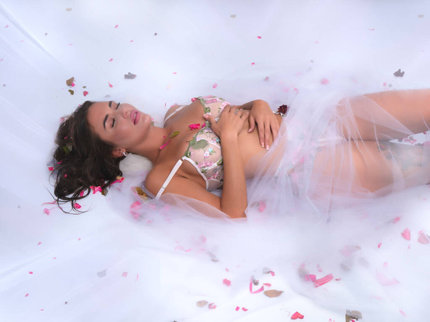 Alto ángulo de sensual joven hembra en lencería acostada en cómoda cama cubierta con velo transparente y pétalos rosados - Foto, Imagen