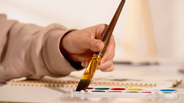 Cropped view of woman holding paintbrush near watercolor paint and blurred sketchbook  - 写真・画像