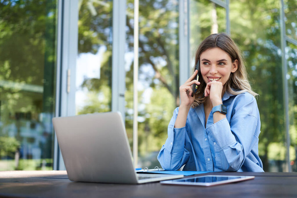 Портрет продавщицы, работающей на свежем воздухе, возле офисного здания. Улыбающаяся корпоративная женщина с ноутбуком, делающая телефонный звонок. - Фото, изображение
