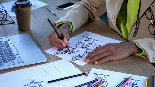 スタジオでラップトップとコーヒーの近くにスケッチを描くアフリカ系アメリカ人のデザイナーの作物のビュー  - 写真・画像