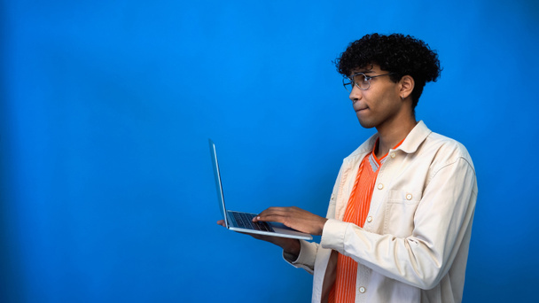 Pensive african american freelancer in eyeglasses using laptop on blue background  - Fotó, kép