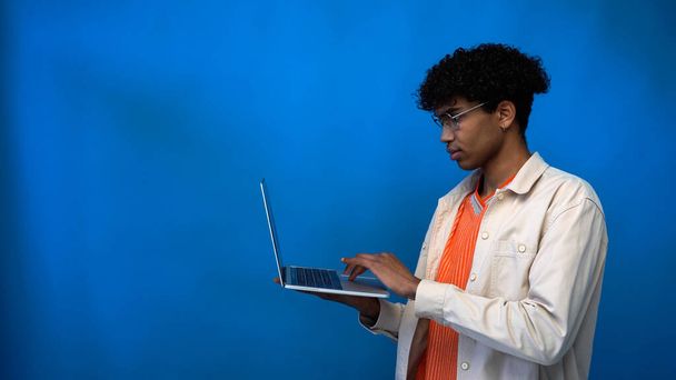 Jonge Afrikaanse Amerikaanse man in bril met behulp van laptop op blauwe achtergrond met kopieerruimte - Foto, afbeelding