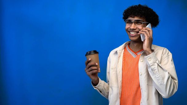 Cheerful african american man in eyeglasses talking on cellphone and holding coffee to go on blue background  - 写真・画像