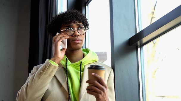 African american businessman in eyeglasses holding coffee to go and talking on smartphone near window in office  - 写真・画像