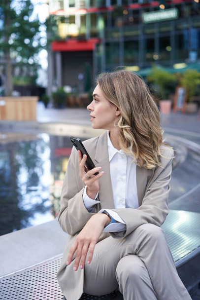 Вертикальний знімок бізнес-леді, дивлячись навколо, тримає мобільний телефон, чекає на місці зустрічі, сидить біля фонтану в центрі міста
. - Фото, зображення